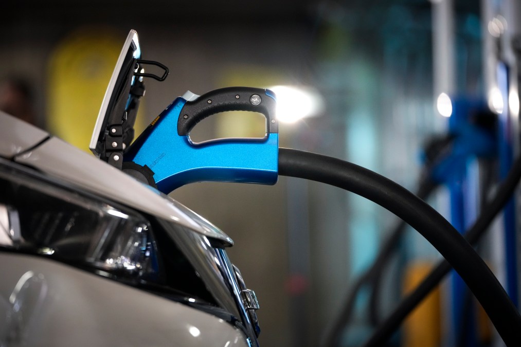 A blue charger is plugged into the front of an electric vehicle. Only the nose of white the vehicle is shown in the frame.