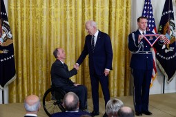 Rory A. Cooper, who uses a wheelchair, shakes hands with President Biden in the East Room of the White House. A uniformed man stands next to them holding a National Medal for Technology and Innovation to be presented to Cooper.