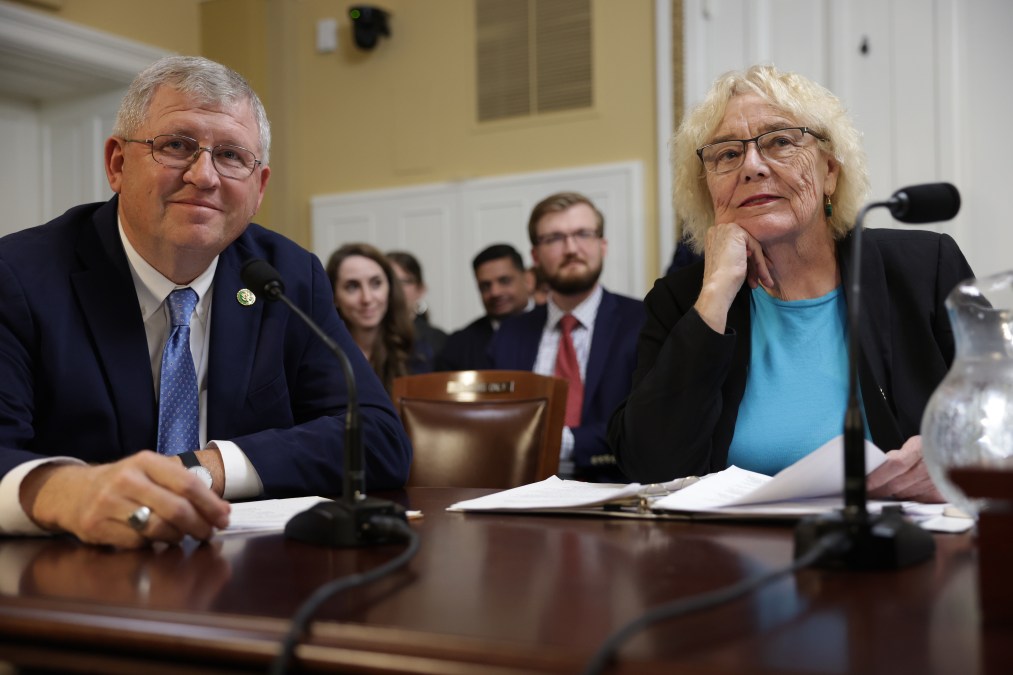 Reps. Zoe Lofgren, D-Calif., and Frank Lucas, R-Okla. sit at a dark wooden table with microphones and papers atop it in a House hearing room.