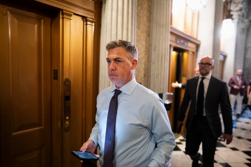 A politician in a dress shirt and tie walks with his phone in his hand.