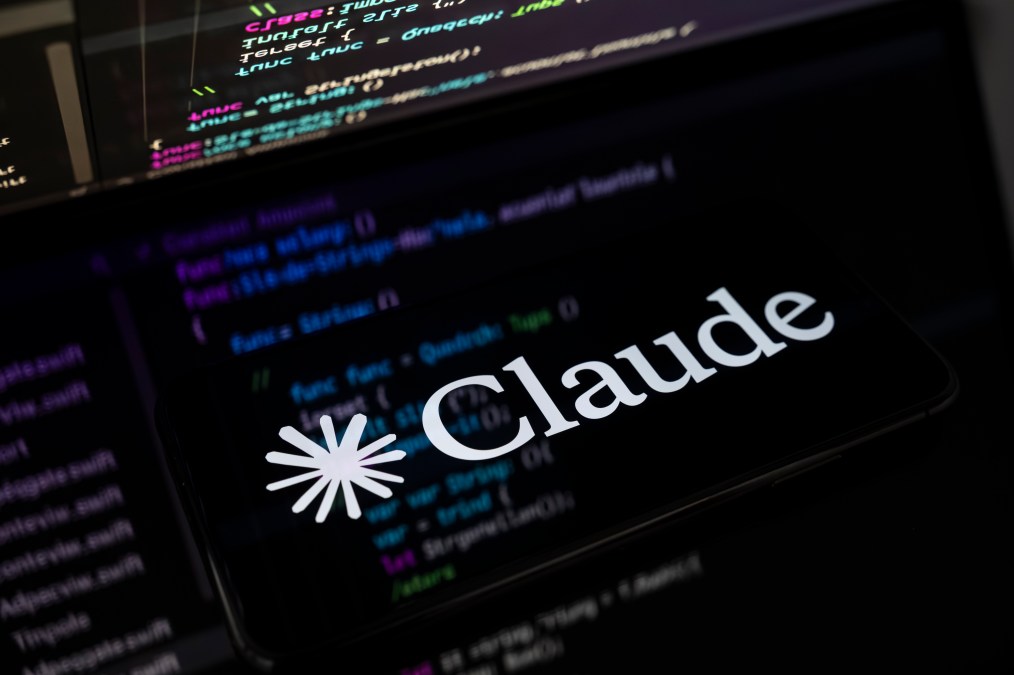 The Claude AI logo is displayed on the screen of a smartphone placed on a reflective surface onto which lines of computer code are projected.