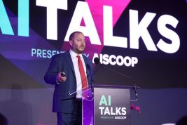 Chase Ausley stands on stage behind a lecturn with the words "AI Talks" written on it in large block letters. A matching AITalks backdrop fills the stage behind him. He is wearing a navy suit with a red tie.