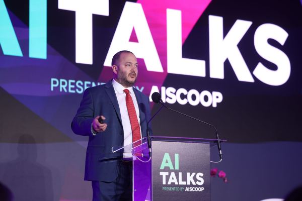 Chase Ausley stands on stage behind a lecturn with the words "AI Talks" written on it in large block letters. A matching AITalks backdrop fills the stage behind him. He is wearing a navy suit with a red tie.