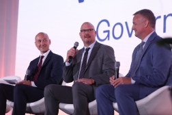 Three men in suits speak on stage into a microphone
