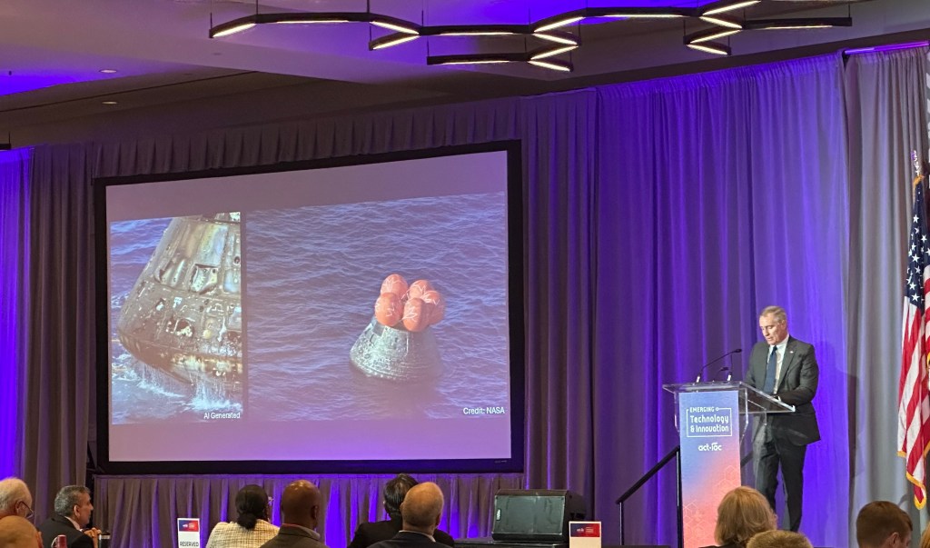 Troy LeBlanc delivers remarks onstage from behind a lectern. To his right, a screen displays a side-by-side comparison of an AI-generated image and real image of the Orion capsule for the Artemis II mission. The AI image shows heavy damage, including scorches and holes. The real image shows the capsule apparently unscathed.