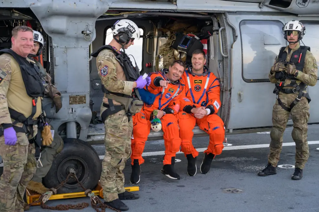 The two astronauts sit on the edge of the landed helicopter. They're both wearing bright orange jumpsuits and smiling. Wiseman points at the camera.