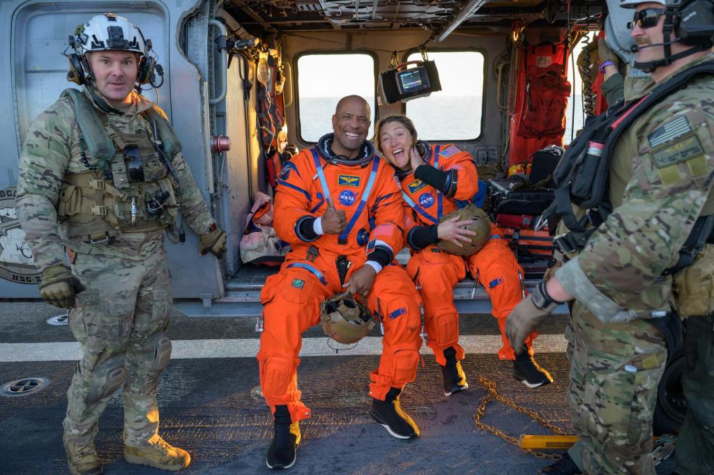 Glover and Koch sit on the edge of a landed helicopter and smile for the camera. Glover holds a "thumbs up" and Koch leans in. They both wear bright orange NASA jumpsuits and hold their helmets in their laps.