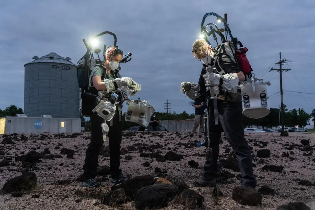 Young and Caswell are shown in a moon-like rock yard at JSC in Houston. They both wear medical face masks, large light-color gloves and carry large backpacks with rods and lights sticking out of the top. It appears to be dusk and the lights illuminate their work. White bags hang at their hips. They are focused on their task.
