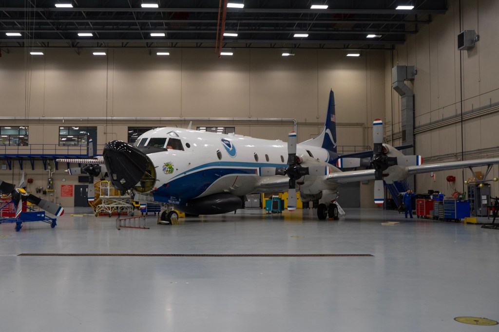 A WP-3D Orion aircraft is parked at an angle in a large aircraft hangar. The plane is white with two shades of blue swooshes on its side. It has four propellers on its wings and a spear-like probe covered in red and white stripes that sticks directly forward and extends past the nose of the plane on its left side. The nose, painted black, is lifted as it awaits maintenance.