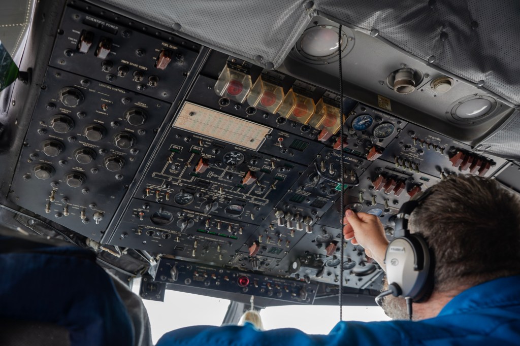The photo is from a vantage point in a seat behind a NOAA pilot. The camera points up over his left shoulder toward a panel of controls on the ceiling. The panel is black and studded with dozens of buttons, knobs and switches of varying shapes and sizes. The panel shows signs of age, including clear covers that on a few buttons starting to yellow.