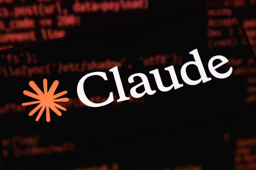 The image shows the Claude logo on a black screen. The logo is a star-like burst of orange followed by the word "Claude" in white text. Code appears reflected on the surface of the screen.